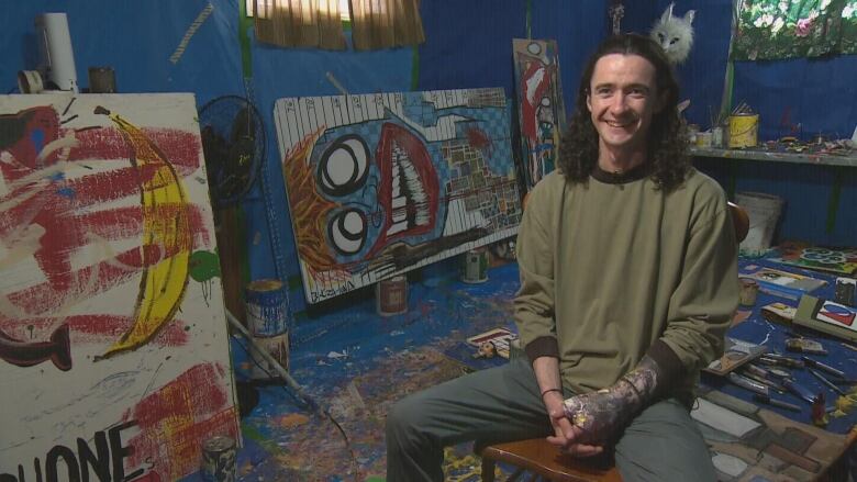 A man sitting on a chair, smiling at the camera with artwork and paint behind him.