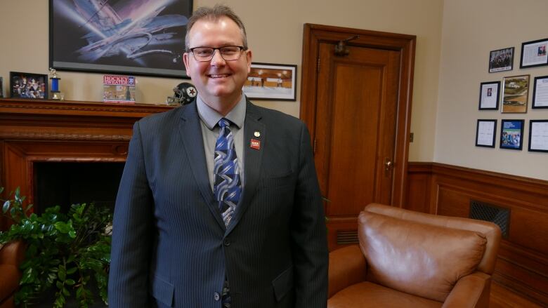 A man in a suit wearing glasses smiles in an office.