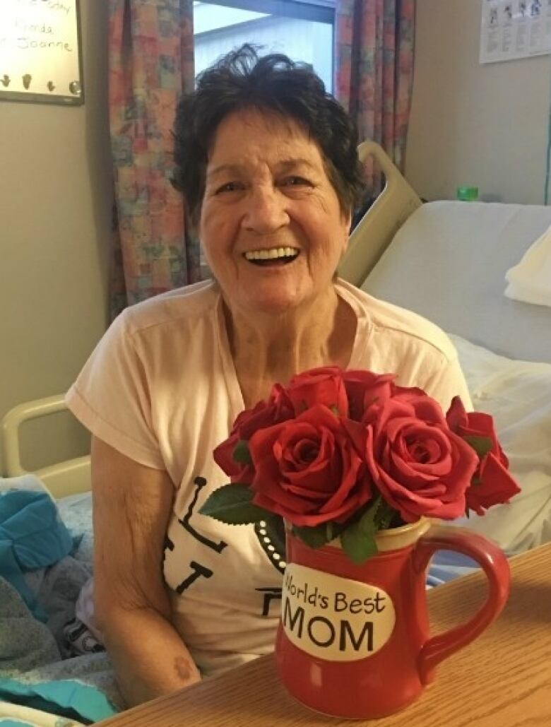 An old woman laughs. She sits on a hospital bed. In front of her on a table is a mug with the words 