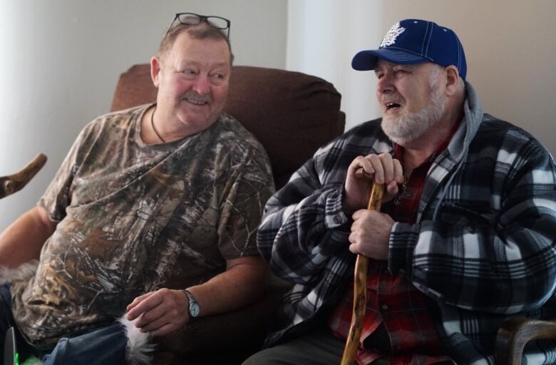 Two men sit in chairs, one looks at the other with a smile. Both have walking canes. 