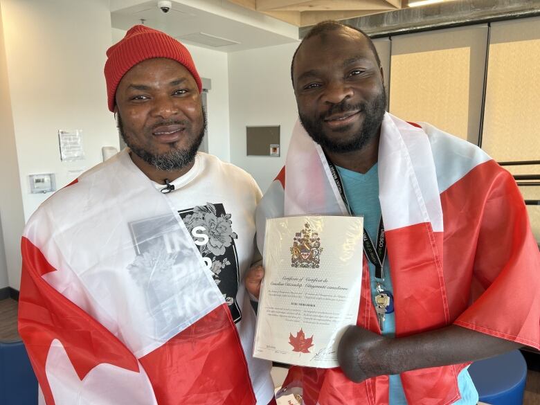 Razak Iyal, left, and Seidu Mohammed pose together for a photo. Mohammed is holding his certificate of Canadian citizenship, while they're both draped with Canadian flags behind their back. 