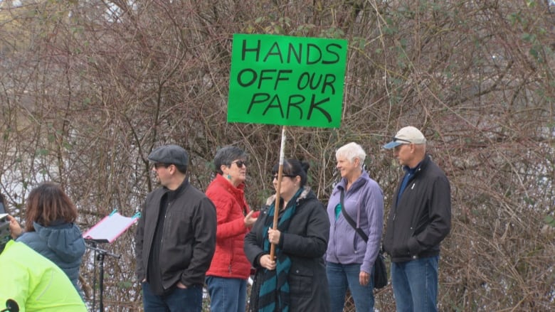 Five people stand at a park in Burnaby. One holds a bright green protest sign that reads 