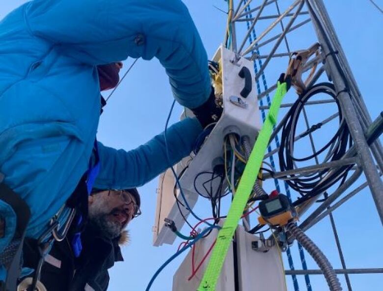 Person in blue coat is working in an electrical box attached to a metal structure. In the background, a man can be seen looking into the same box.