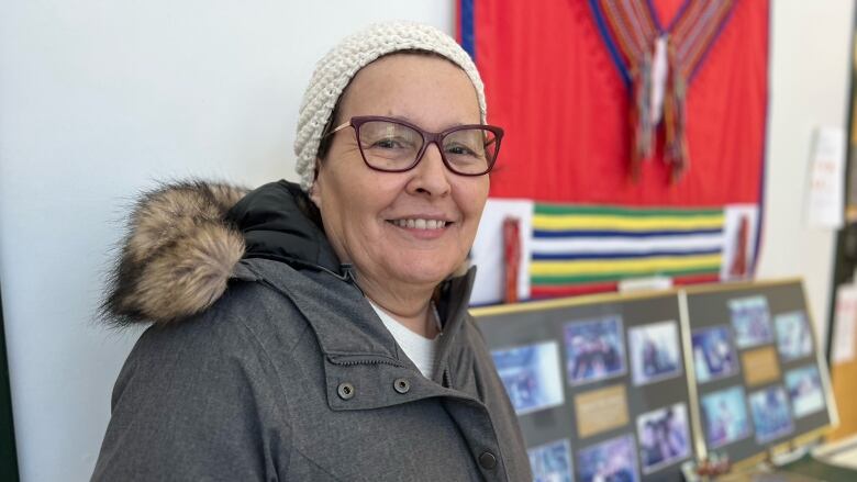 A woman in a winter jacket stands in front of a display of photographs and books. In the background is a decorated wall-hanging.