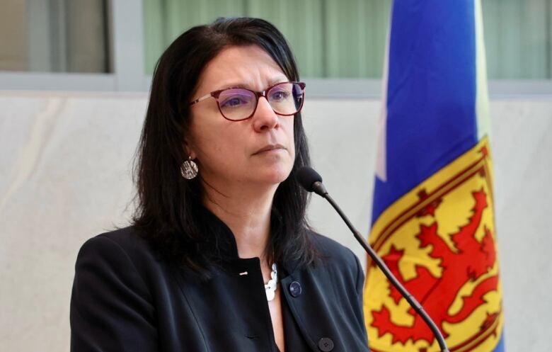 A woman with shoulder-length dark brown hair and glasses and silver earrings and a silver necklace wears a black jacket. She is standing next to a Nova Scotia flag. 