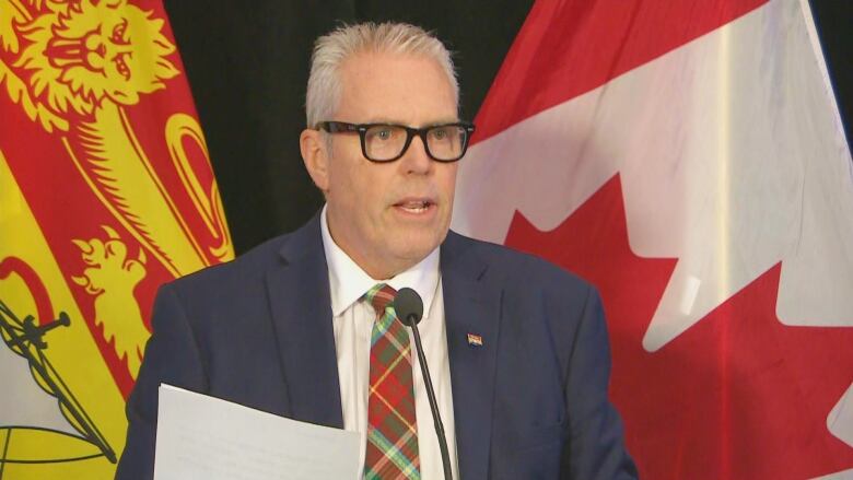 A man wearing a suit and a plaid tie standing in front of a New Brunswick flag, left, and a Canadian flag, right.