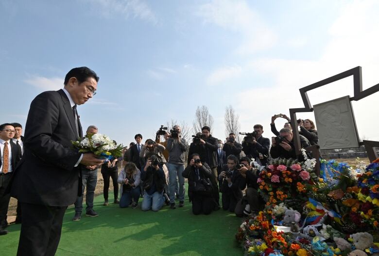 A man in a suit is shown holding flowers and reflecting before a memorial, with dozens of observers in the background.