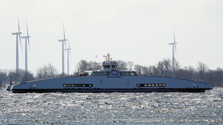 A white ferry with a Canadian flag flying from its cabin can be seen floating on the water. Trees and wind turbines can be seen on an island behind it.