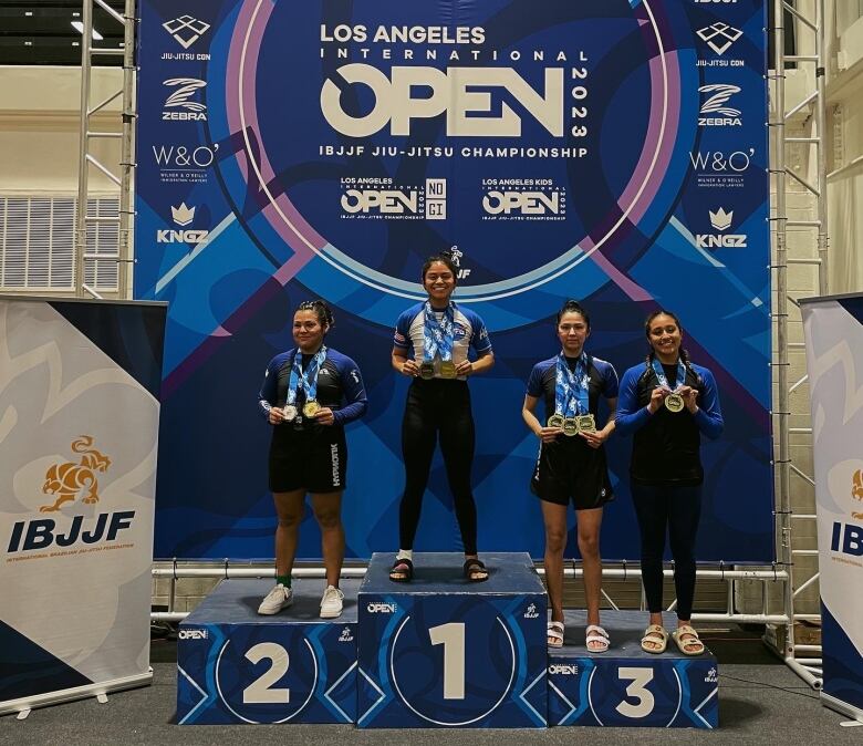 Four women stand on a tournament podium: one on the first place stand, one on the second place stand and two on the third place stand. 