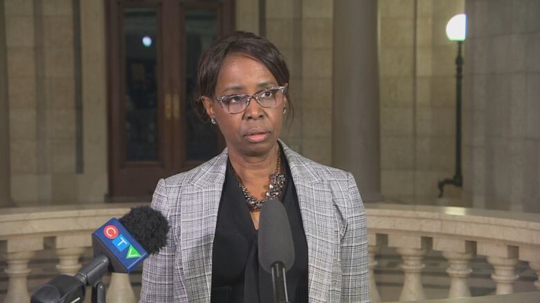 A woman wearing glasses and a blazer with her hair pulled back speaks in front of microphones.