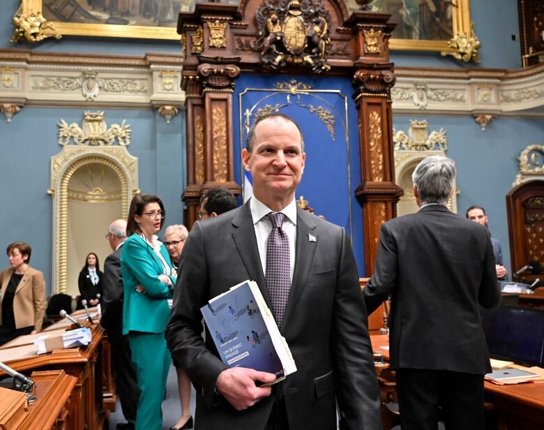 Man in a suit with a book walks into a legislature room with lots of gold trim on walls.