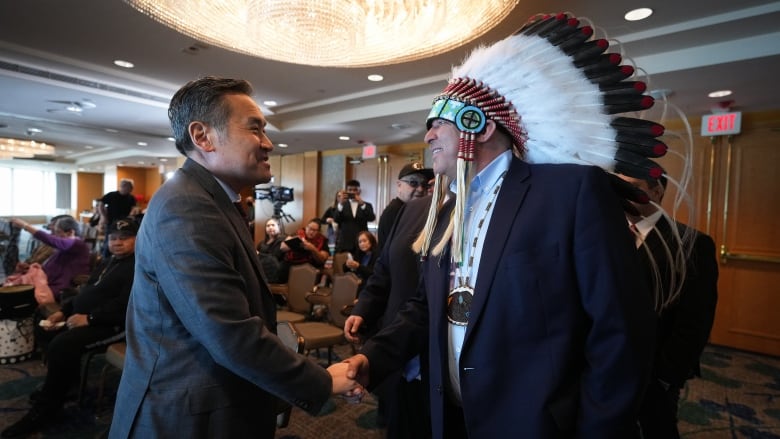 A man in an Indigenous headdress shakes hands with another man in a suit under a conference room chandelier.