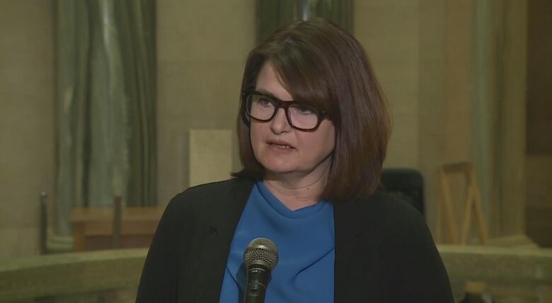 A woman wearing glasses, a blue shirt and black blazer stands behind a microphone on a stand inside the rotunda of the Saskatchewan Legislative Building. 