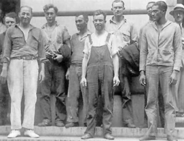 A black-and-white photos shows a group of men wearing old clothing standing together, smiling at the camera.