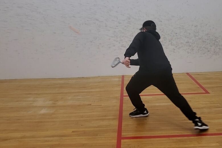 A teenager wearing a black hoody, black pants and a black baseball hat hits a squash ball. The player is partially sighted and is playing squash for the first time.