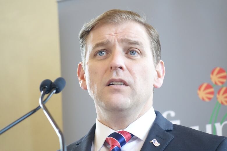 A man wearing a blue suit standing behind a podium with two microphones. The pitcher plant of the Newfoundland and Labrador logo is visible behind him. 