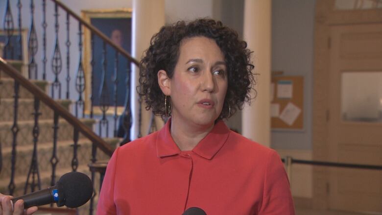 A woman with short brown curly hair wears an orange blazer. She is standing in front of a staircase.