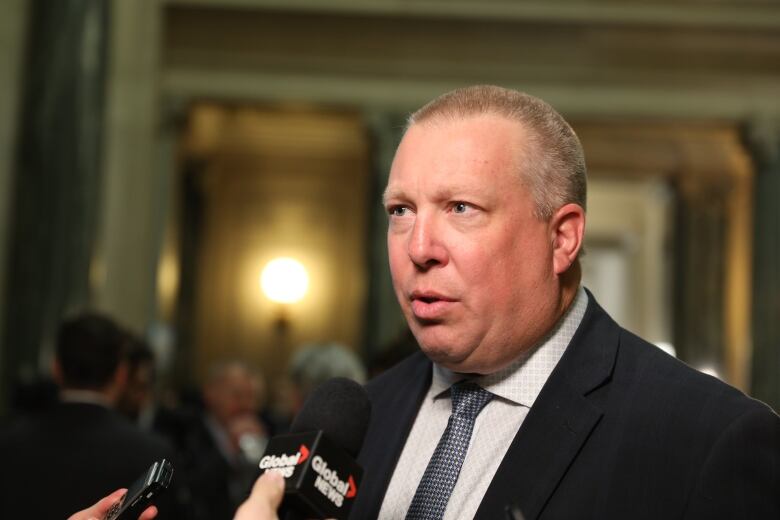 A white man with a buzz cut is wearing a suit with a blue-and-silver tie. He is speaking into a microphone, while standing in a rotunda.