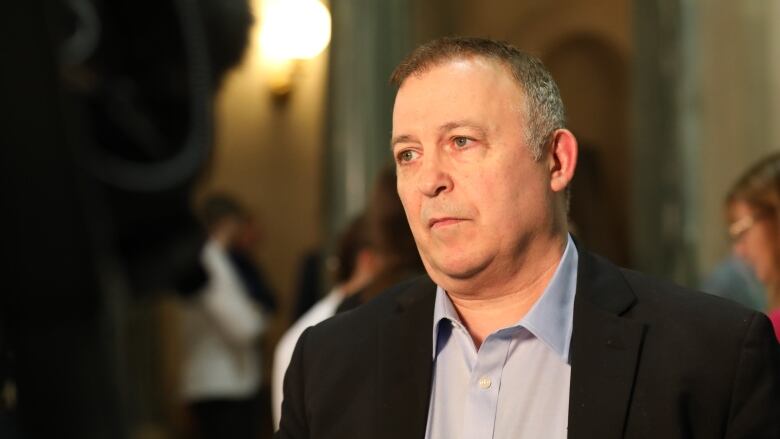 A white man, with short greying hair, is wearing a black blazer over a light blue dress shirt, the top button of which is undone. He is standing in rotunda. There are people standing about in the background.