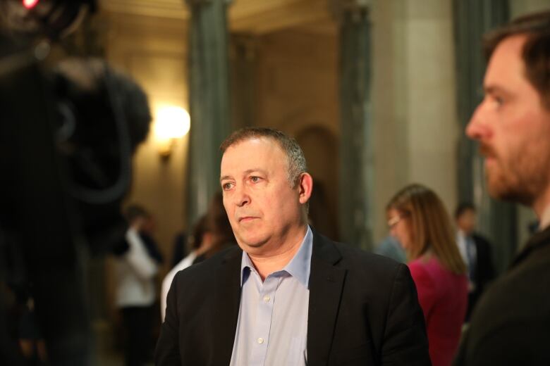 A white man, with short greying hair, is wearing a black blazer over a light blue dress shirt, the top button of which is undone. He is standing in rotunda. There are people standing about in the background.