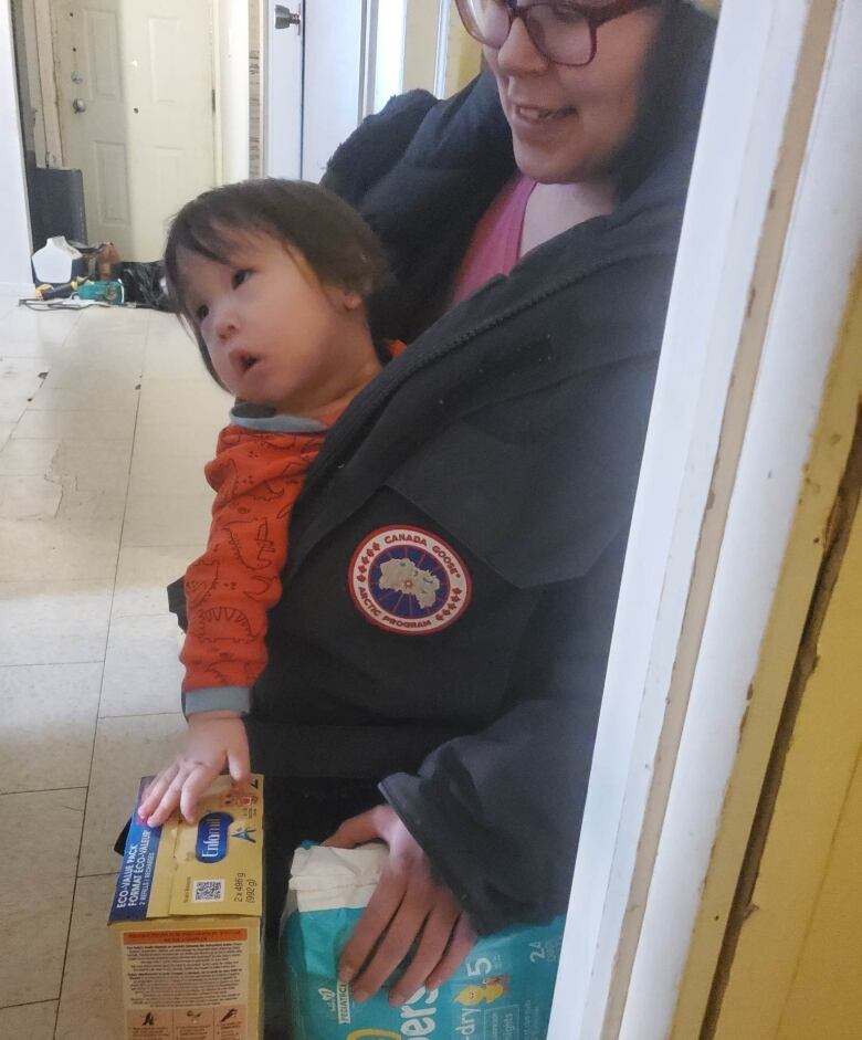 A woman stands carrying a toddler and holding a box of infant formula and a bag of diapers.