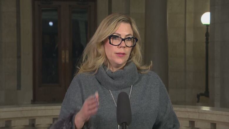 Woman with blond hair and glasses stands at a microphone in the rotunda of the legislature.