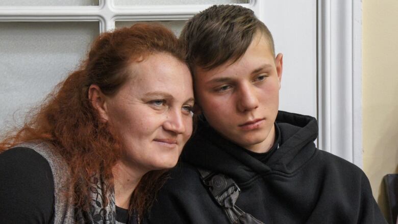 A mother with long red hair smiles and rests against her teenage son, who has a serious expression.