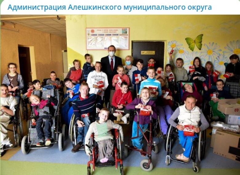 A group of children in wheelchairs in front of a row of adults pose for a photo near a wall painted yellow with a butterfly.