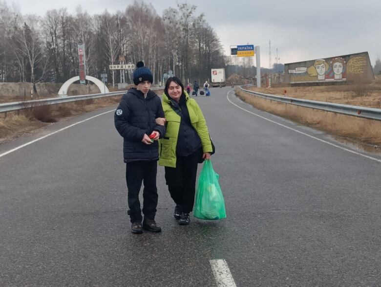 A mother holding a green bag stands next to her young son on an empty road.