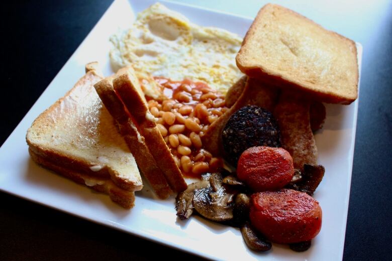 A plate covered in bread, fried eggs, beans, sausage, mushrooms, black pudding, tomatoes and bacon.