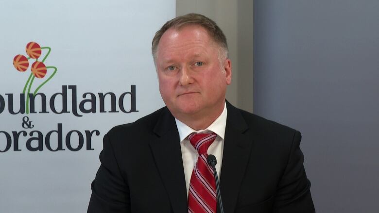 A mean wearing a black suit and red striped tie sits in front of a microphone.