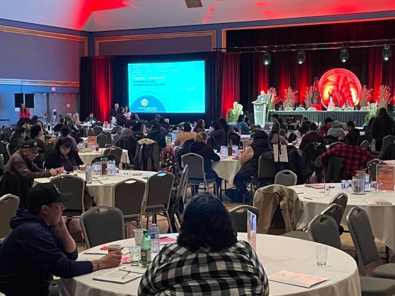 People sit at round tables with white table cloth in front of a big screen for the conference. 