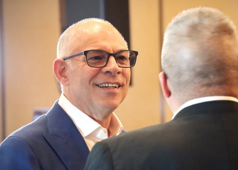 A man with glasses smiles and speaks with someone whose back is to the camera.