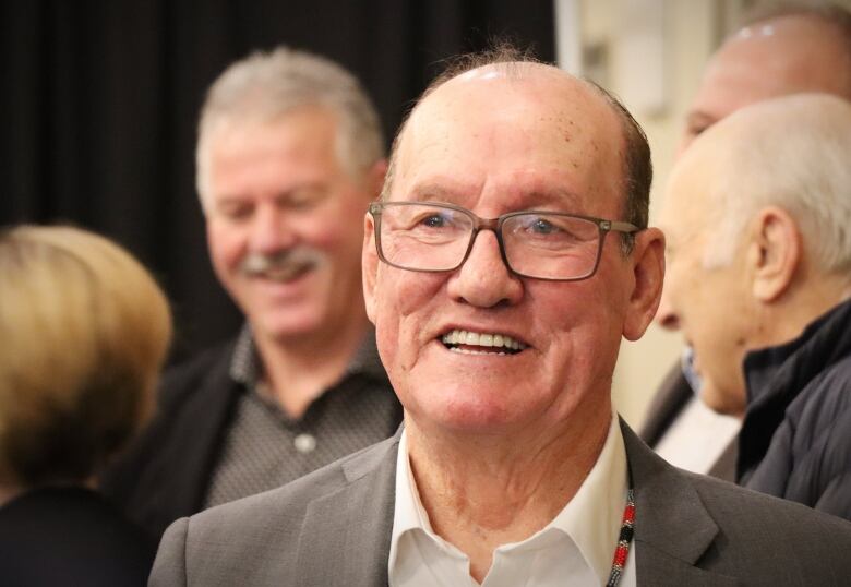 A man with glasses, wearing a grey suit jacket and white shirt open at the collar, smiles.