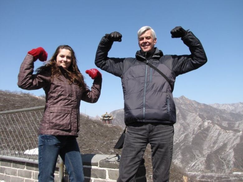 A woman wearing a red mittens and brown winter jacket flexes her arms beside a man with a black winter jacket and black gloves in a matching flexing pose in front of mountains.