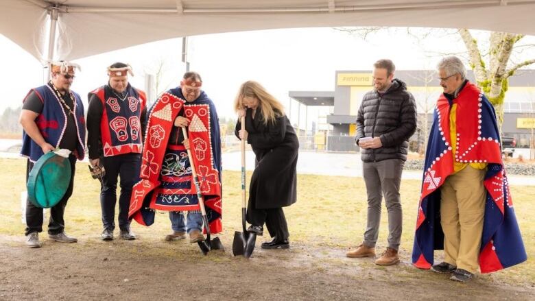 Men wearing traditional Indigenous cloaks and in regular clothes stand to the side as a man wearing an Indigenous cloak and traditional headband shovels dirt with a woman holding her own shovel. 