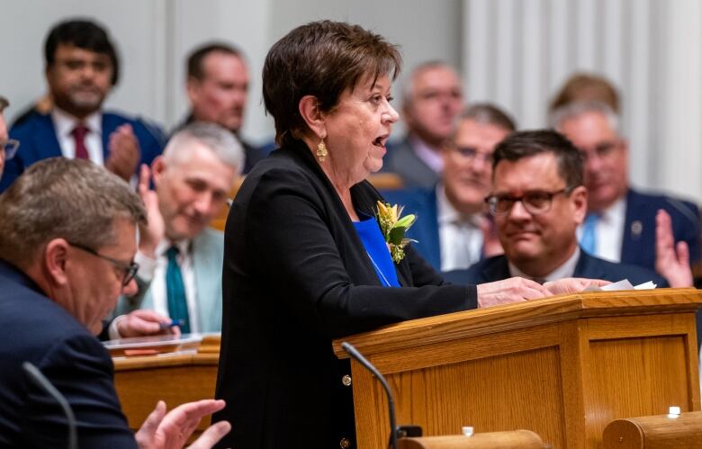 A woman with short brown hair and wearing a black suit speaks while standing at a podium surrounded by a group of men.