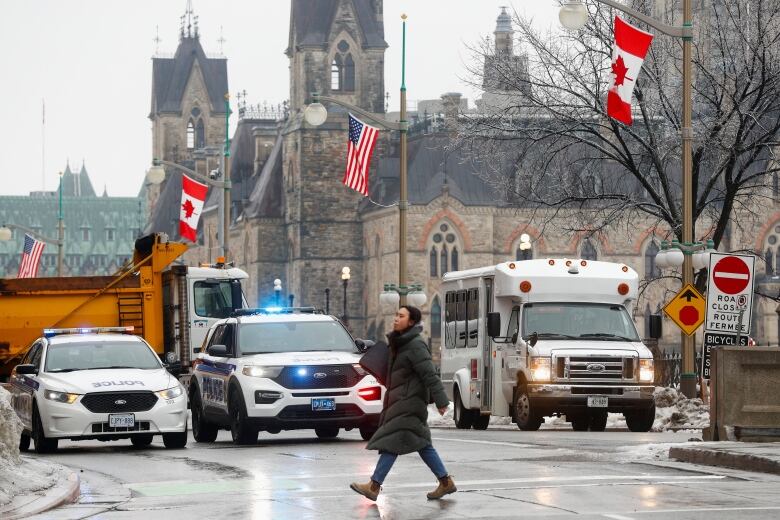 Someone in a winter coat crosses a rainy street. Two police vehicles and a dump truck are in the background.