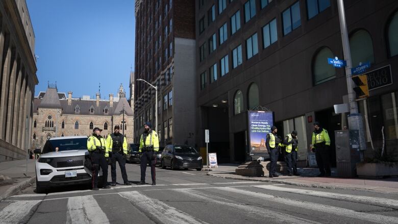 Six police officers in yellow jackets watch a closed street.