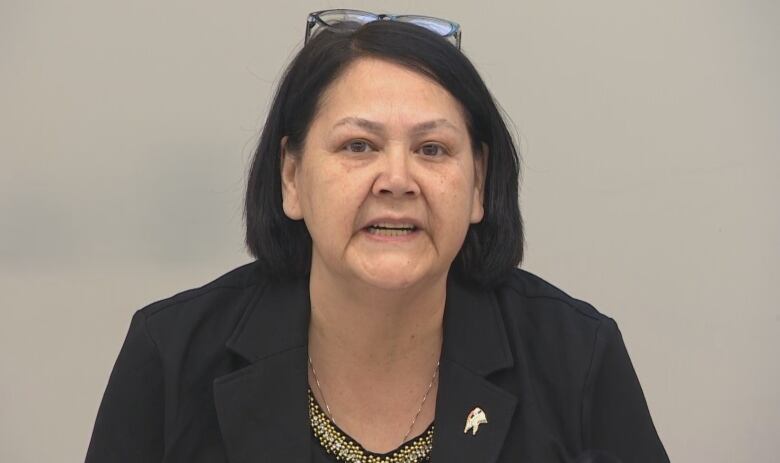 A woman with black hair and eyeglasses set on top of her head, wears a black blazer and with a pin on the lapel.