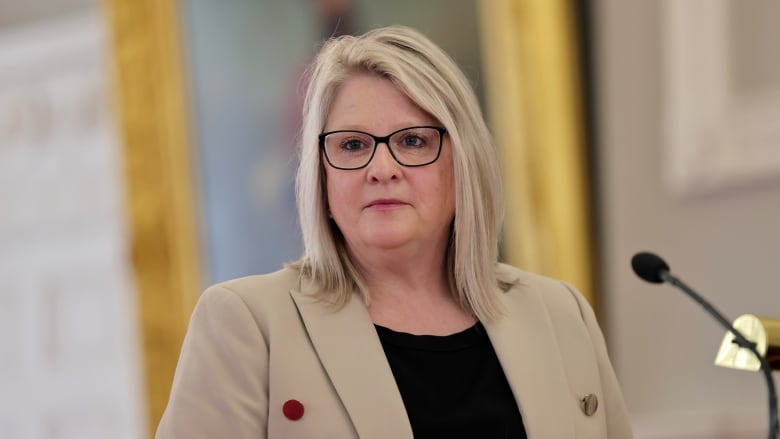 A blonde woman with glasses standing wearing a beige coat. 