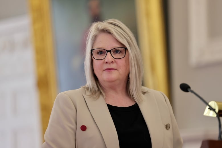 A blonde woman with glasses standing wearing a beige coat. 