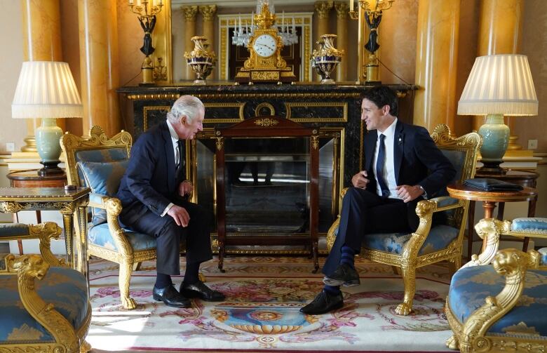 Two people sitting in chairs in front of a fireplace talk with one another.