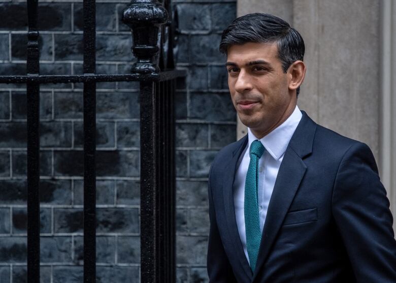 A man with dark hair, wearing a grey suit and green tie, smiles as he leaves a grey-bricked residence.