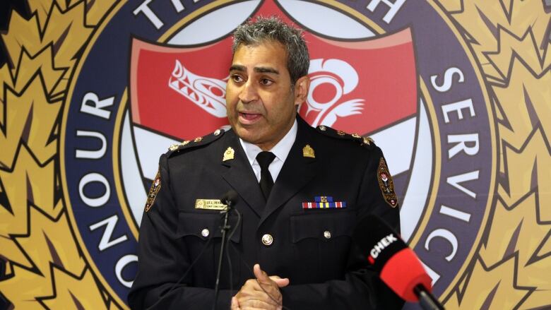 A man in a police uniform stands before a podium speaking with his hands clasped together. Behind him on a screen are the words 'Honour through service.' 