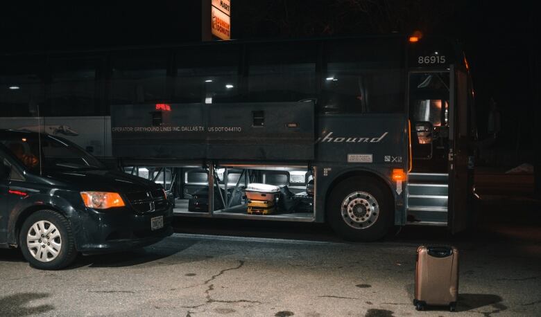 Luggage compartment of the bus is open revealing its content. A lone suitcase stands on the parking lot pavement in front of the bus. 