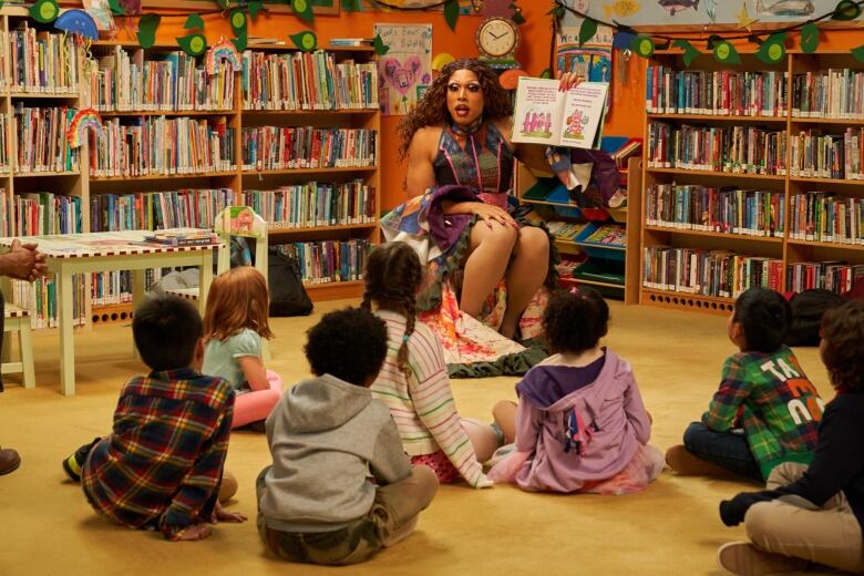 Children sit cross legged on the floor of a library. Seated in front of them a drag queen reads from a book. 