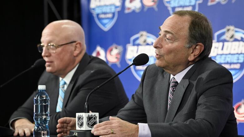 Two men in suits speak in front of a microphone, with a glowing screen with sports logos on it behind them.