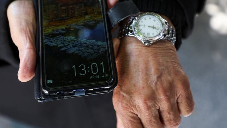 A man holds a smartphone in his right hand, showing the time 13:01, while a watch on his left hand shows the time 12:01.