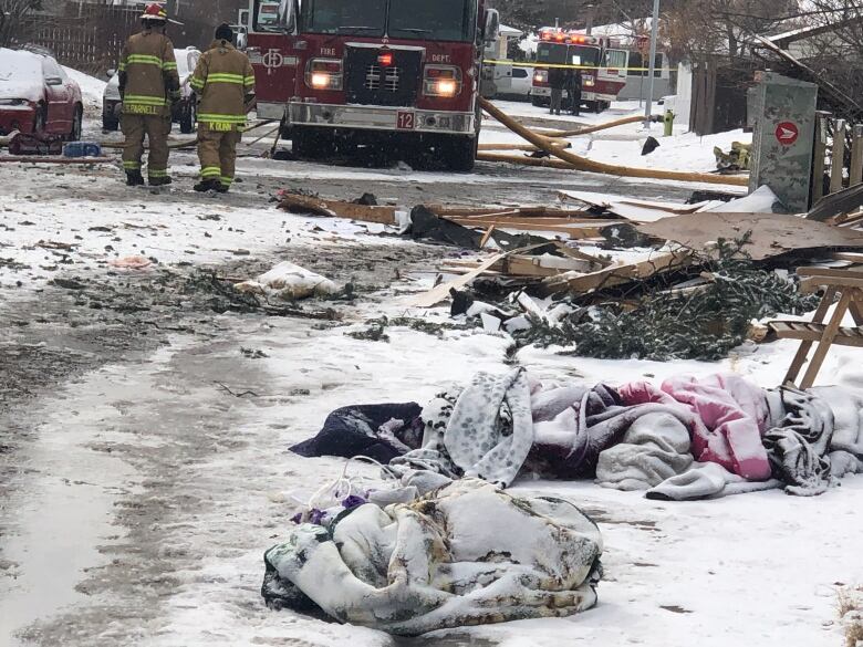 Snow-covered blankets and debris sit on the road while firefighters walk to an engine in the distance. 